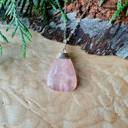 Rose quartz teardrop pendant on a chain against a natural wood and greenery background